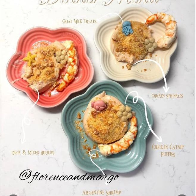 A styled dinner menu board featuring three pet food dishes on flower-shaped plates, labeled Goat Milk Treats, Chicken Sponges, and Duck & Mined Berries with Argentine Shrimp, on a white marble background.